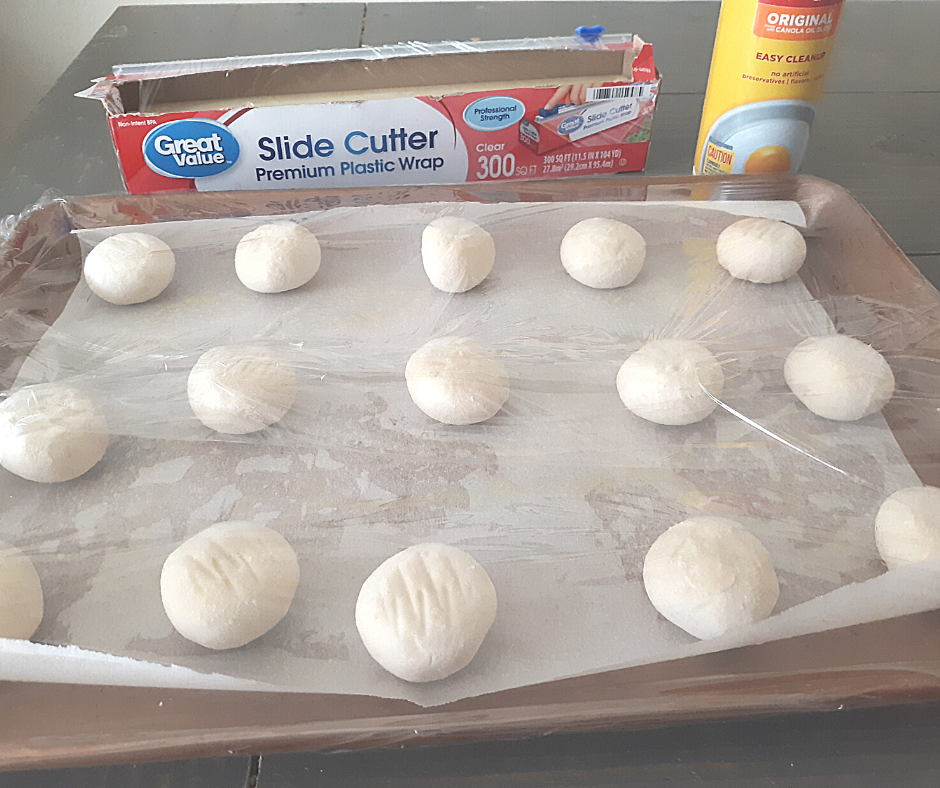 frozen dough balls on parchment paper, on a metal sheet pan, covered in plastic wrap