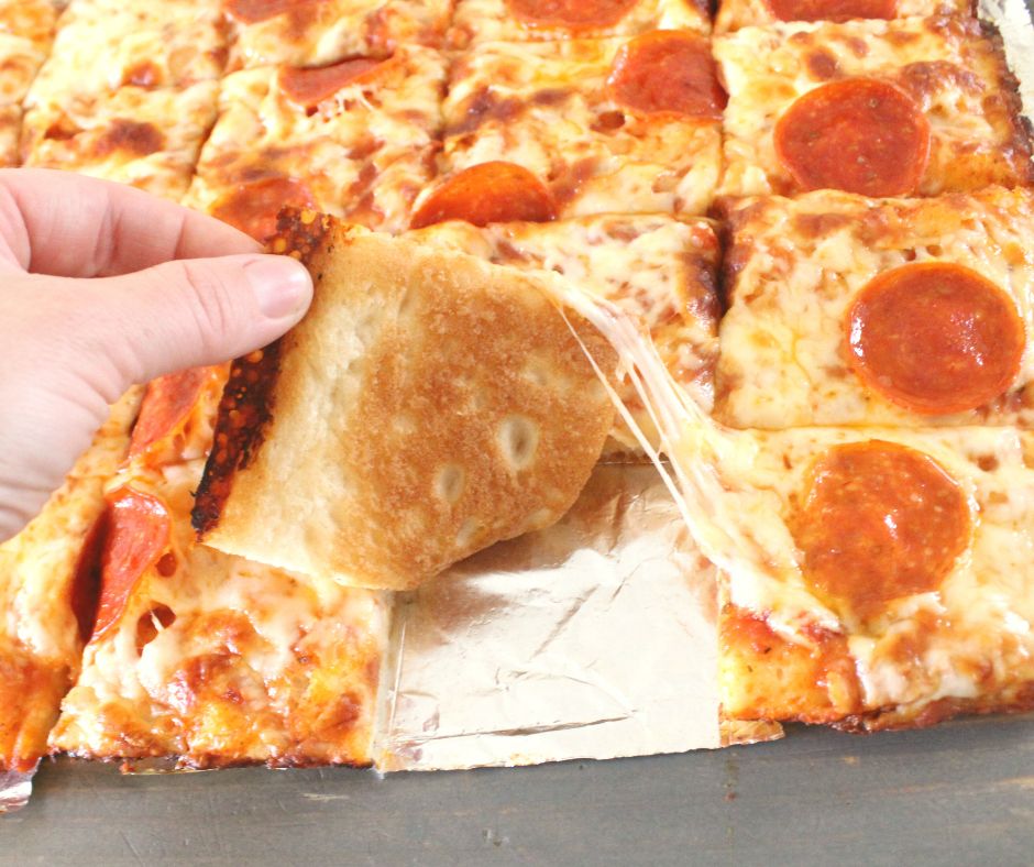 a hand lifting up a square of sheet pan pizza to show it is browned on the crust