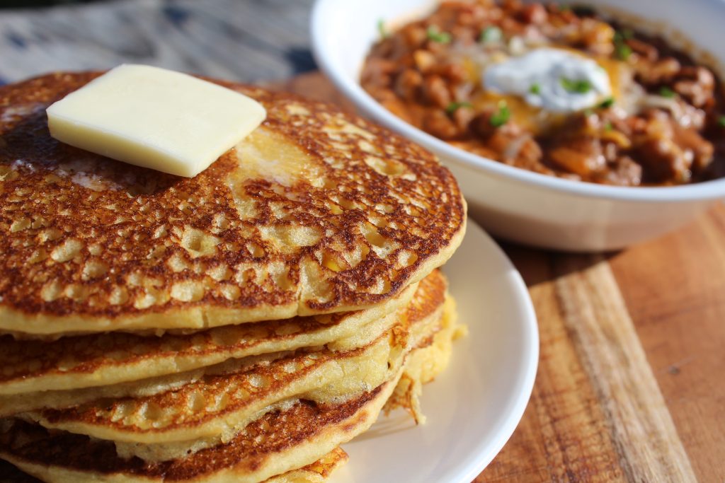 close up of a stack of cornbread pancakes with butter and chili in the background