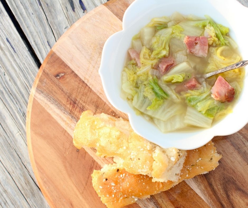 cabbage soup with ham in a white bowl with bread sticks on a wooden cutting board.